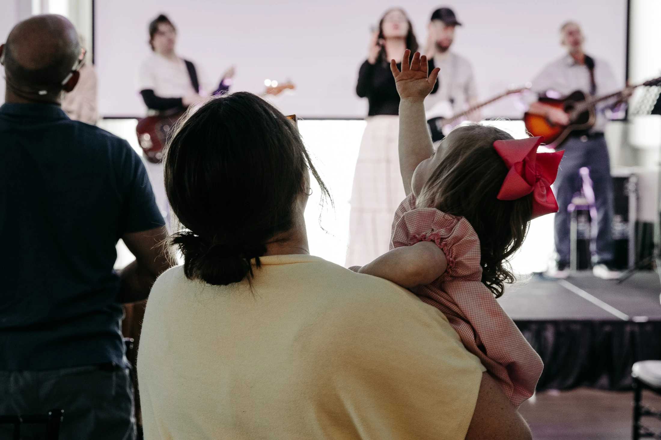 From behind, a woman holds a small child with a pink bow who is raising her hand while watching a worship band perform on a brightly lit stage.