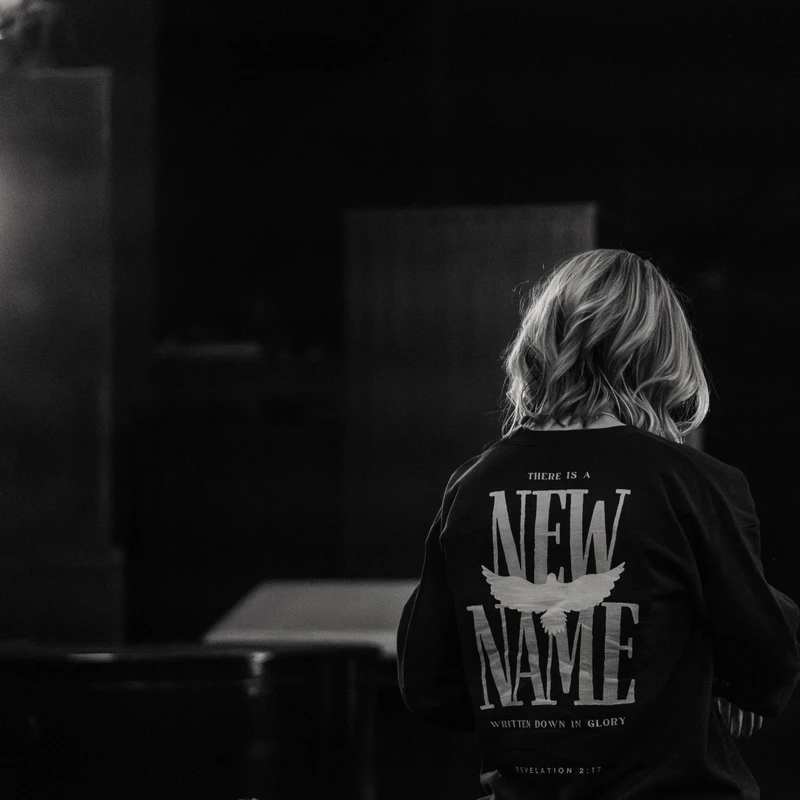 A black and white photo from behind of a person wearing a shirt with text that reads, "There is a new name written down in glory," from Revelation 2:17.