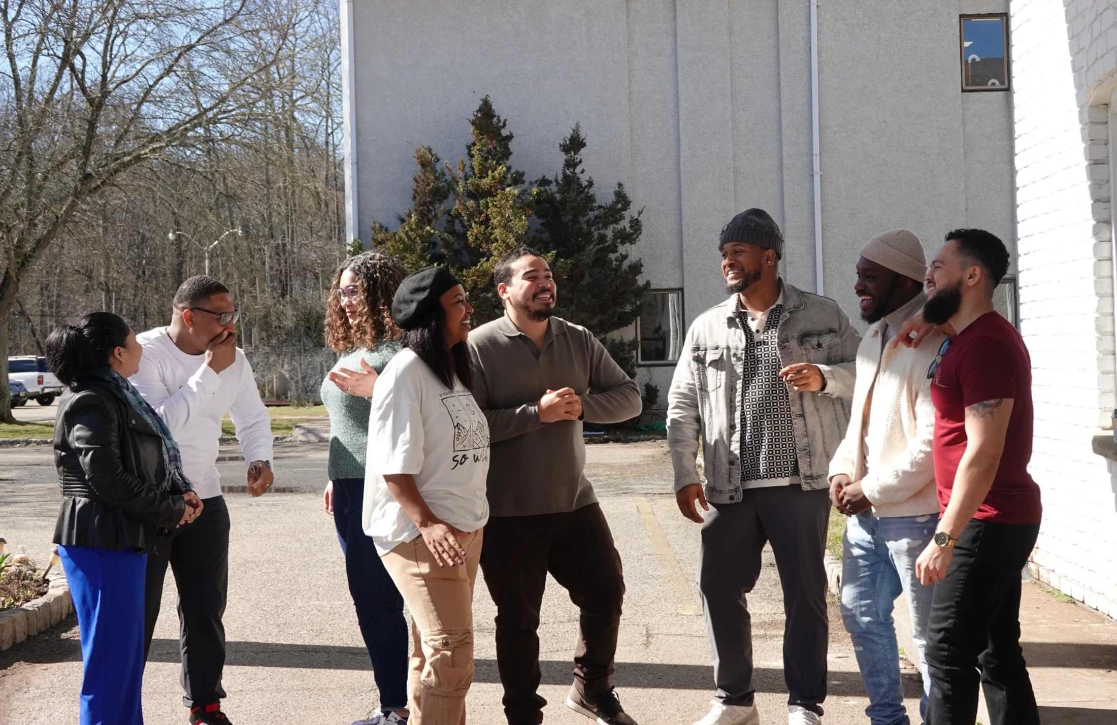 A diverse group of eight adults stand together outside on a sunny day, smiling and laughing while engaged in a pleasant conversation.