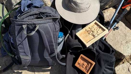An overhead shot of hiking gear, including a backpack, sun hat, and trekking poles, laid out on sunlit rocks with commemorative wooden mountain plaques.