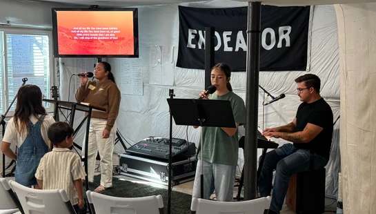 A worship band with two female singers and a male keyboardist performs in a room. A screen shows lyrics, and a small boy watches from the front row.
