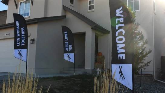 Three black feather flags with Christian messages like 'WELCOME' and 'JESUS LOVES YOU' stand in the front yard of a two-story beige house.
