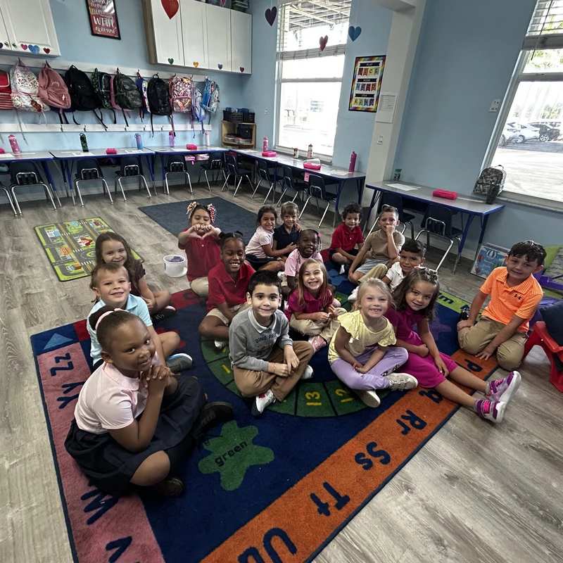 A diverse group of young students sit cross-legged on a colorful alphabet rug, smiling for a group photo inside their bright, well-lit classroom.