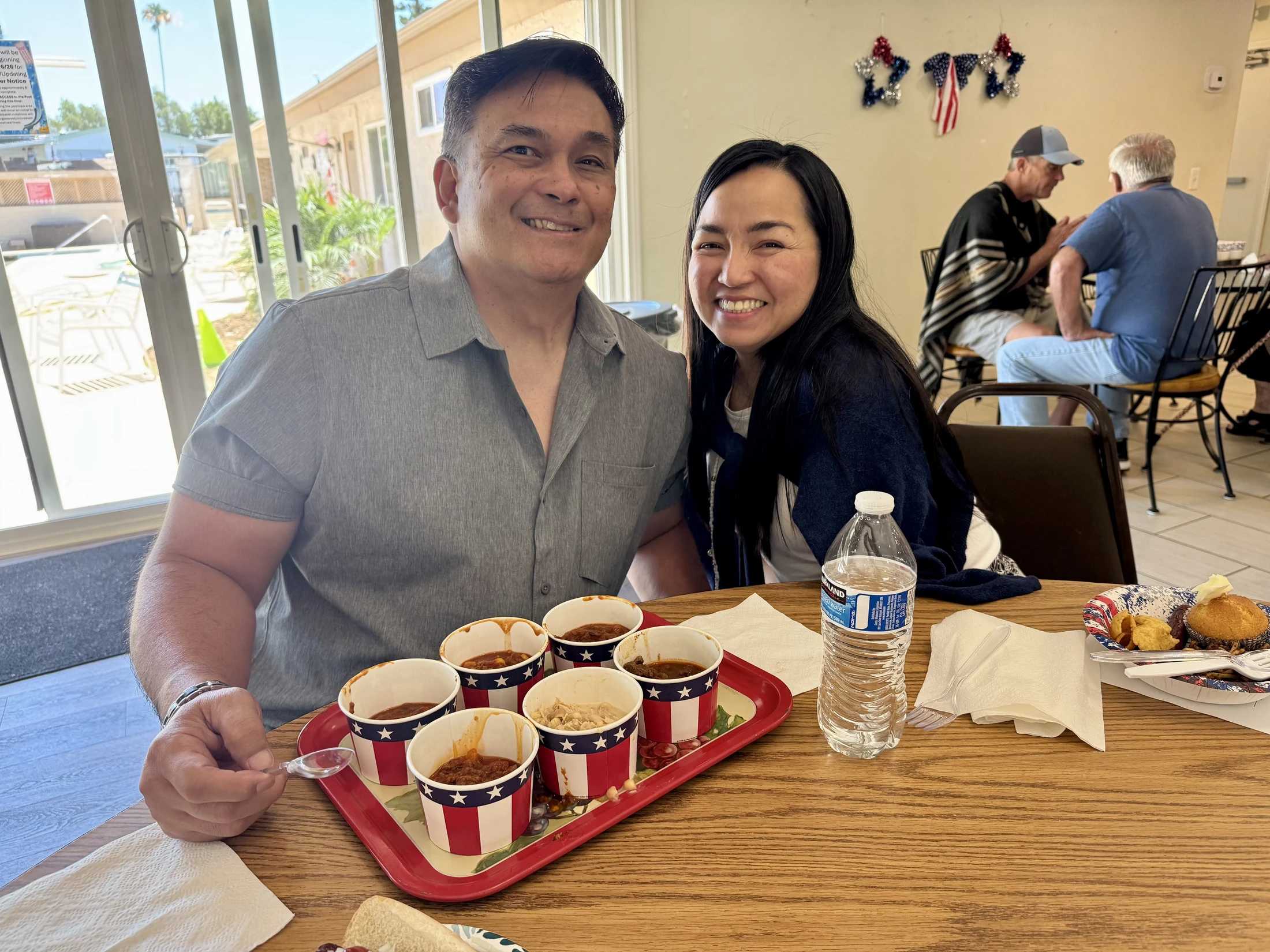 A smiling man and woman sit at a table. The man holds a tray with several small cups of chili decorated with American flags for a tasting event.