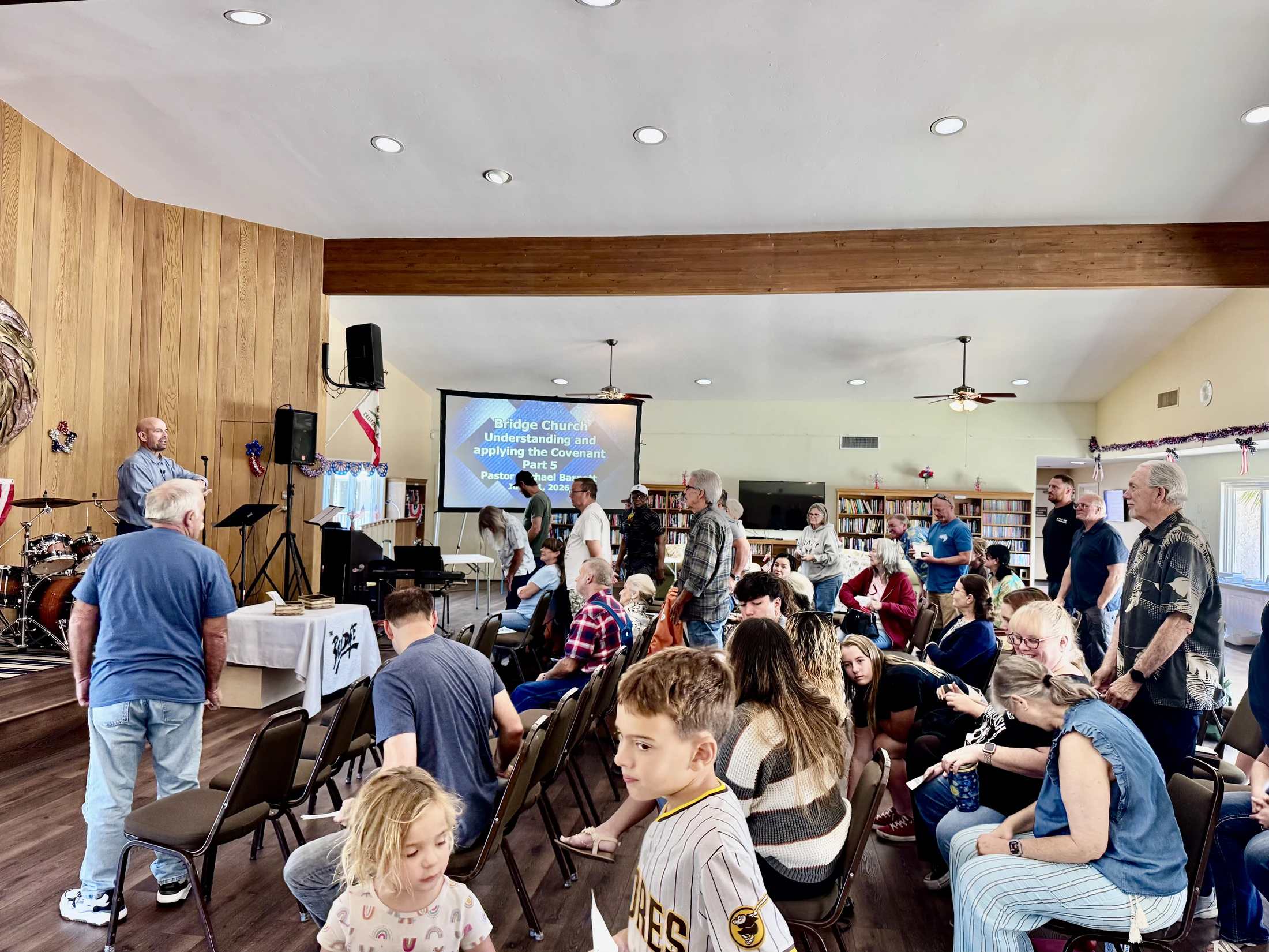 A diverse congregation of people, including adults and children, are gathered inside a hall for a church service, with a pastor speaking at the front.