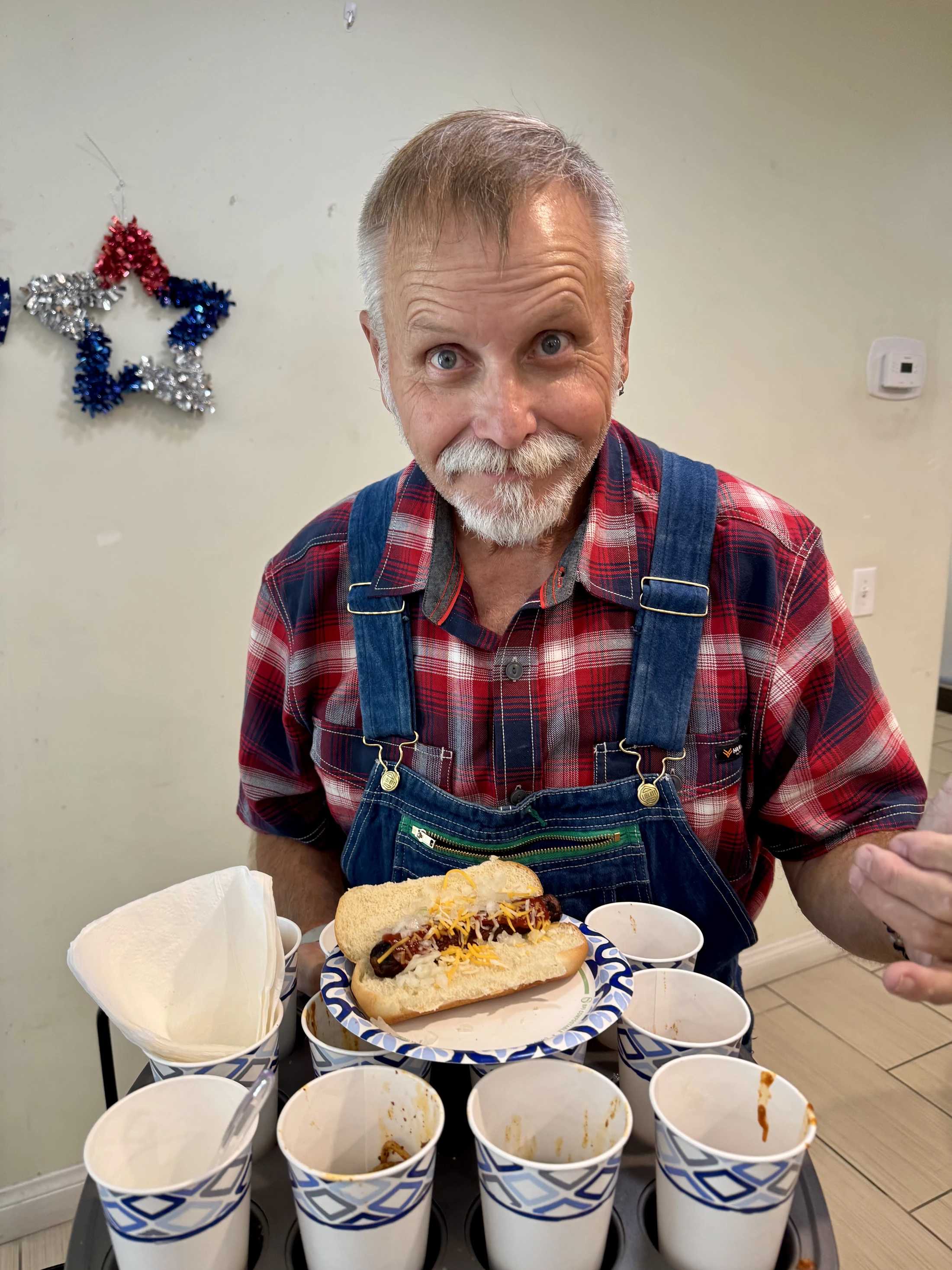 A friendly man with a white mustache wearing a plaid shirt and overalls holds a tray with a chili cheese dog and several small cups for a chili tasting.