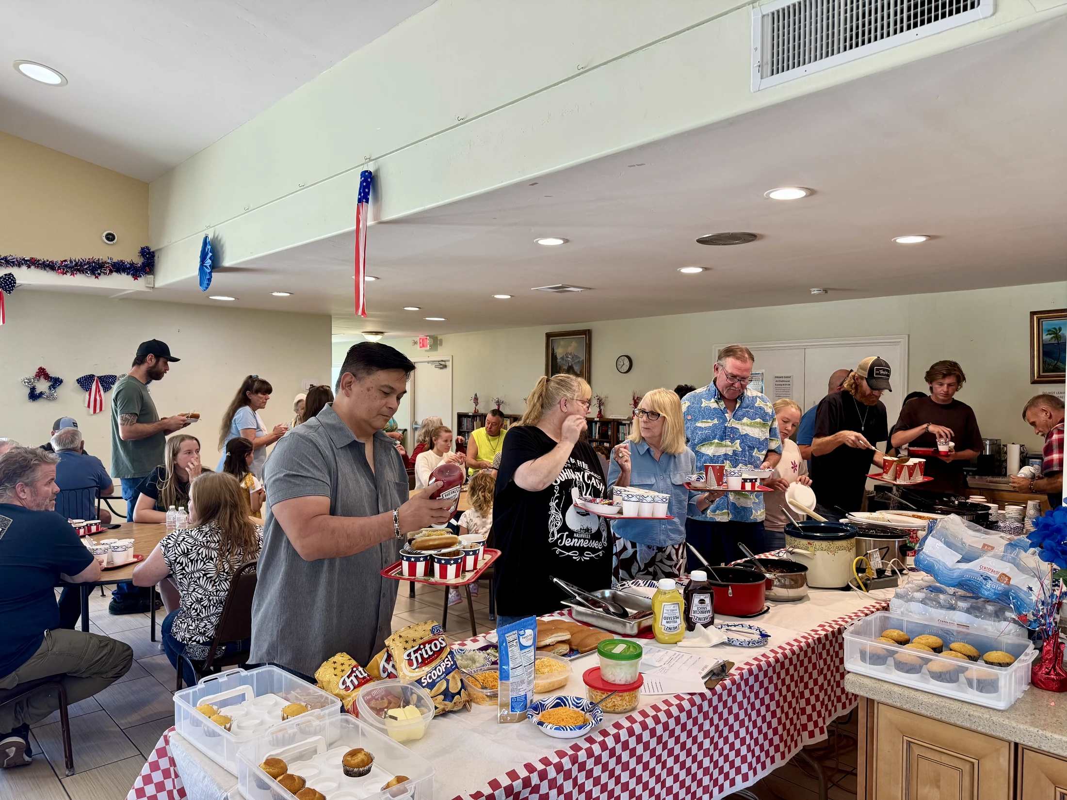 A diverse group of people serve themselves food from a long buffet table during an indoor gathering with red, white, and blue decorations.
