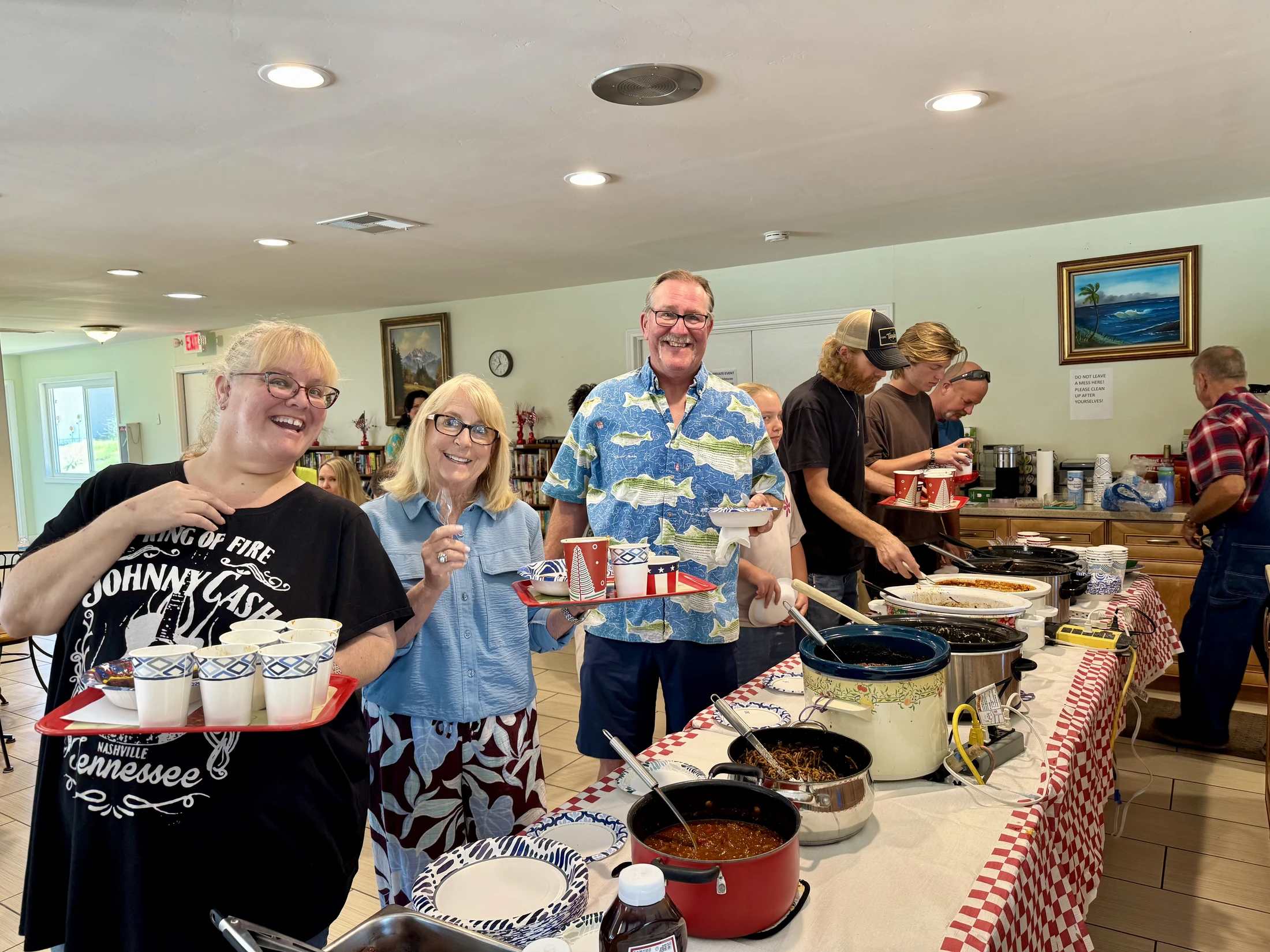 A group of happy people line up at a buffet table filled with crock pots and serving dishes, holding trays with bowls and cups at an indoor gathering.