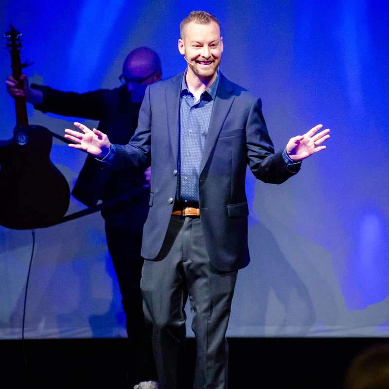 A smiling man in a dark suit stands on a stage, gesturing with his hands. He is lit by blue lights, and a guitarist is visible in the background.