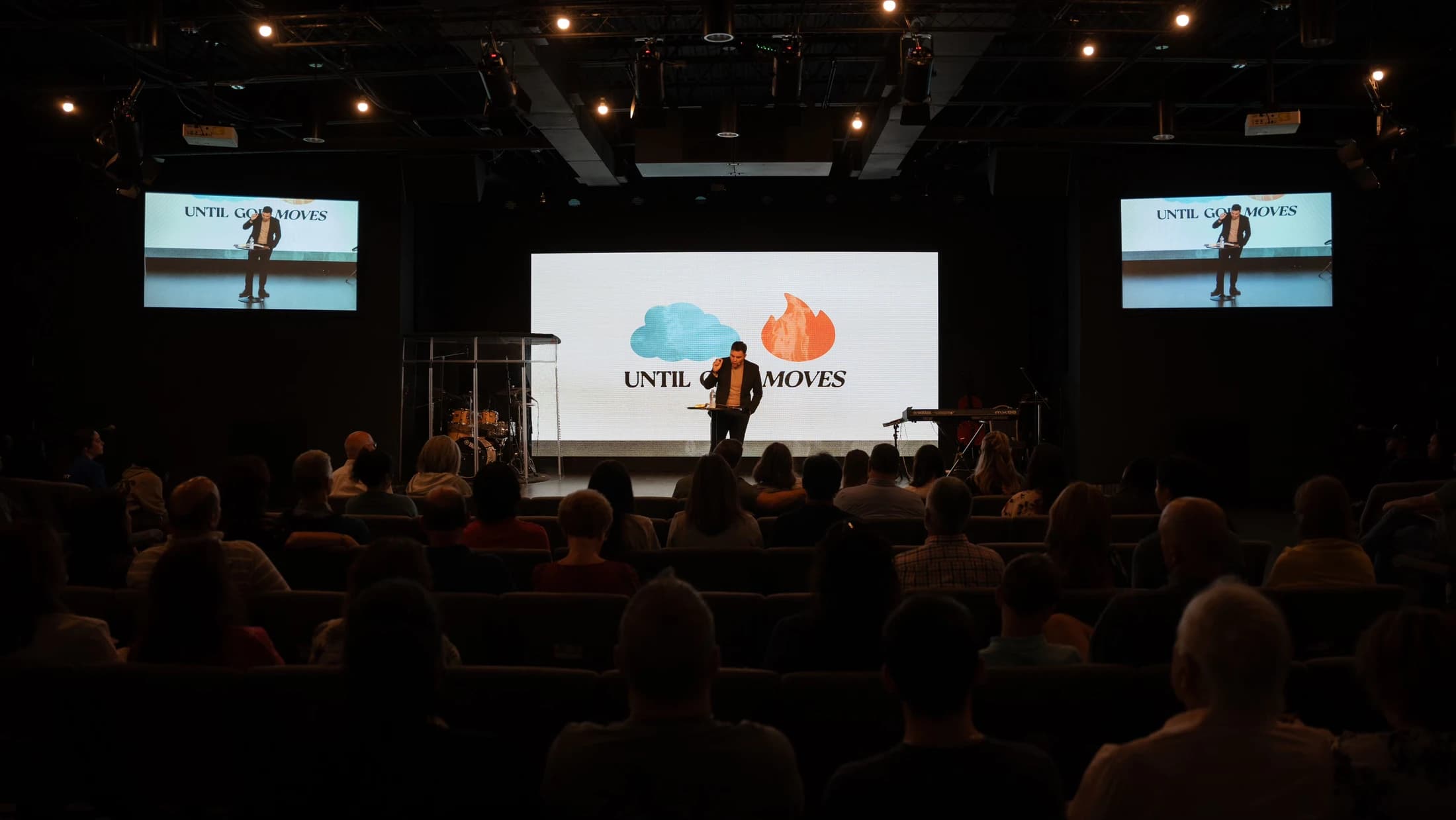 A pastor speaks on a lit stage to a large audience in a dark auditorium. A screen behind him reads 'UNTIL GOD MOVES' between a cloud and a flame graphic.