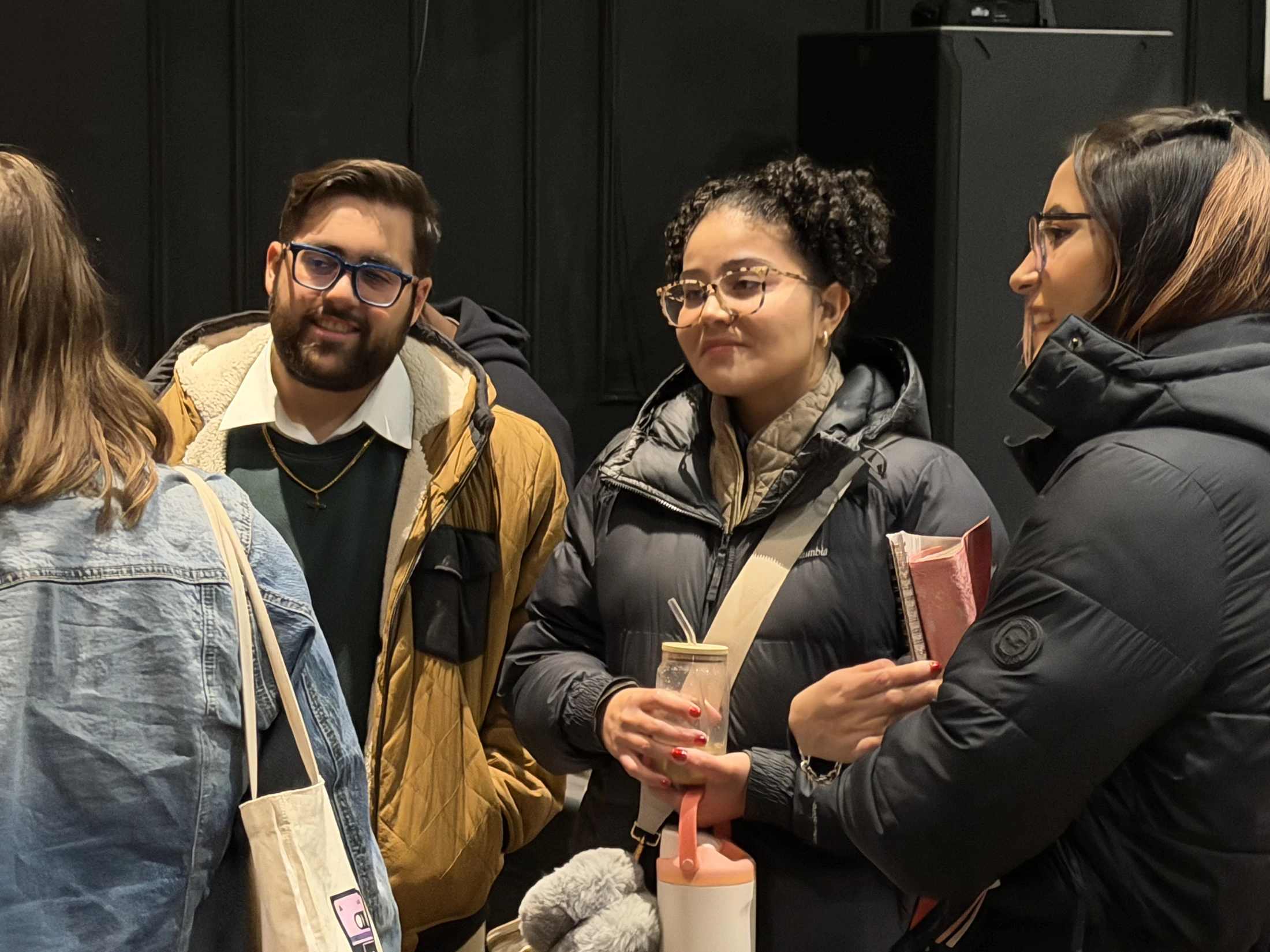 A group of four young adults in casual winter jackets stand indoors against a dark wall, smiling and talking with each other.