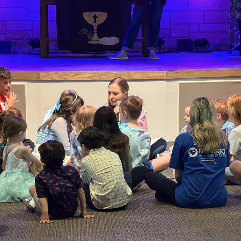 A woman speaks into a microphone while sitting on the floor in a circle with a group of children during a church service. A stage is visible in the background.