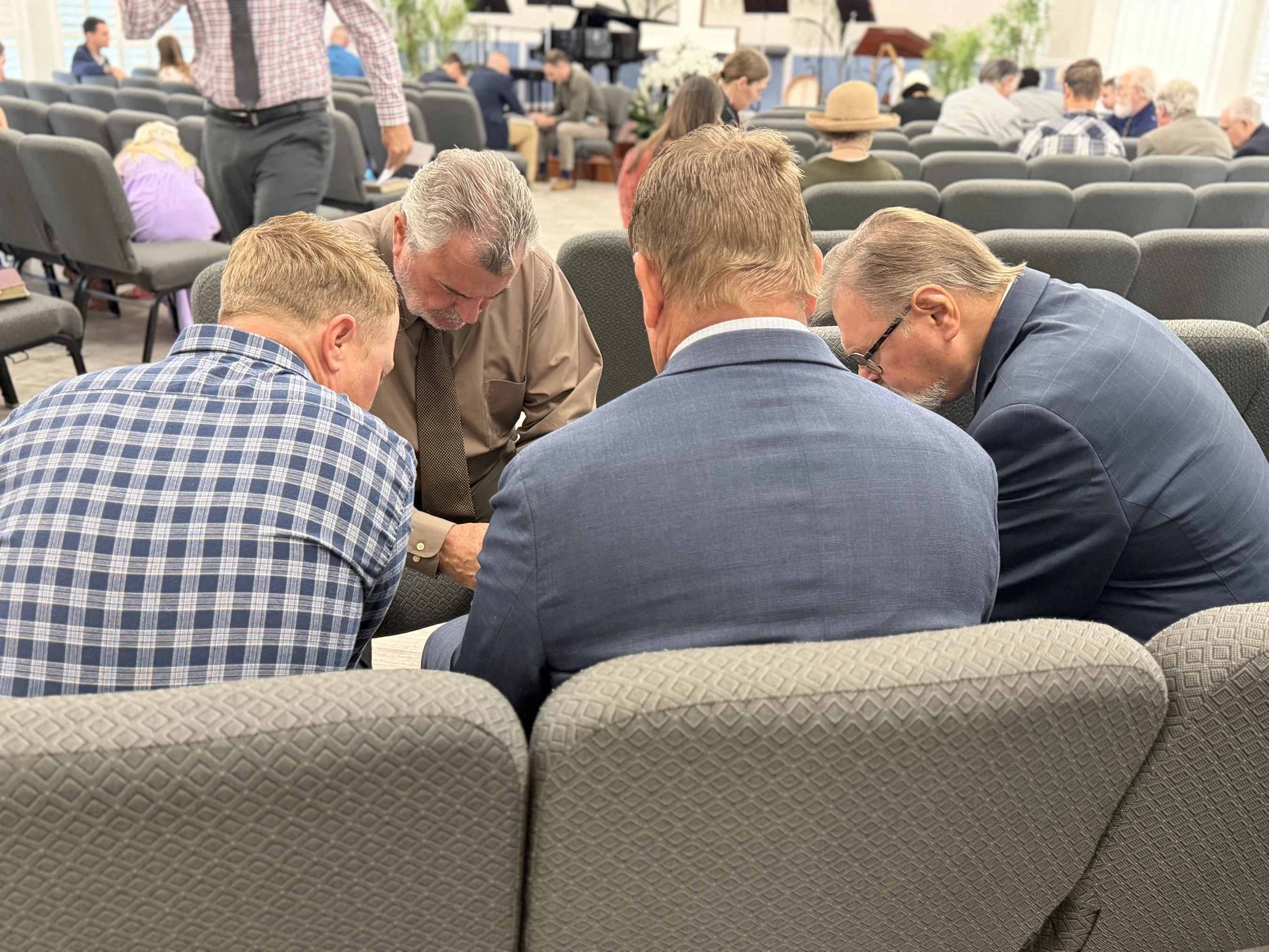 Four men sit huddled together in upholstered chairs inside a large room, bowing their heads in prayer. Other people are seated in rows in the background.