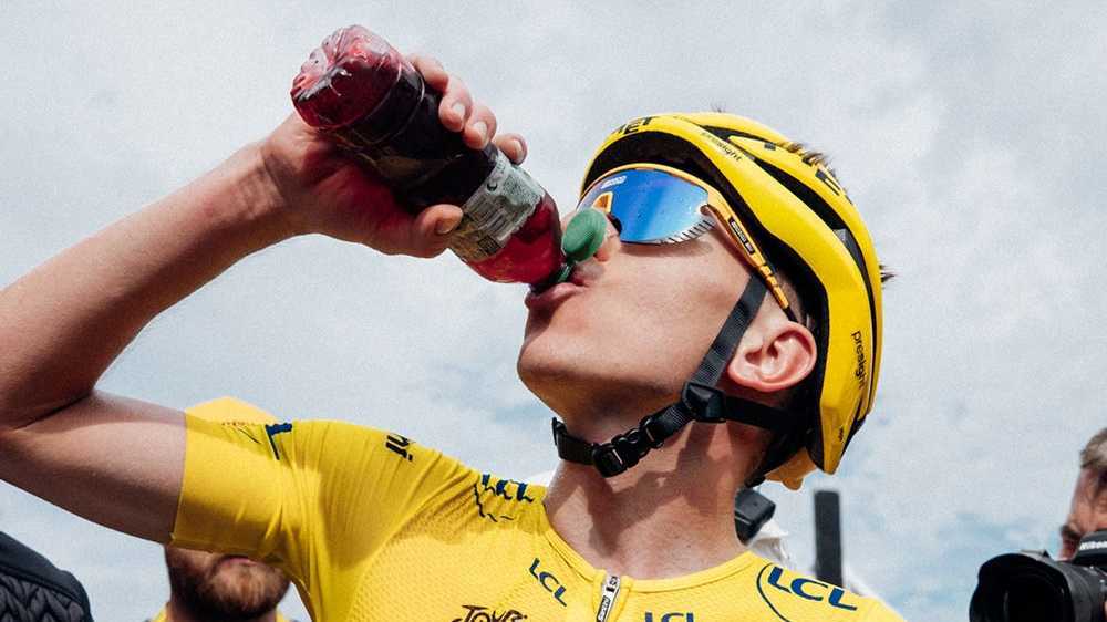 Tour de France champion Tadej Pogačar wearing the yellow jersey drinks Enervit Magic Cherry recovery drink after a stage finish, highlighting post-race recovery and performance nutrition.