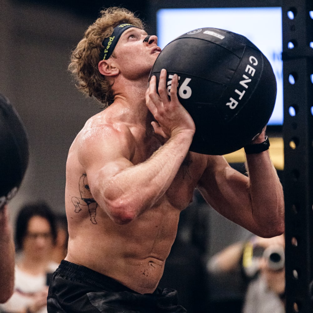 Hunter McIntyre mid-rep during a HYROX workout, shirtless and wearing a “The Feed” headband, lifting a 16kg CENTR wall ball, showcasing strength and focus for BLDR brand lifestyle campaign