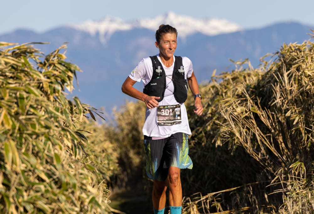Courtney Dauwalter trail running through tall grass with snow-capped mountains of Mt. Fuji in the background, wearing a white shirt and black hydration vest