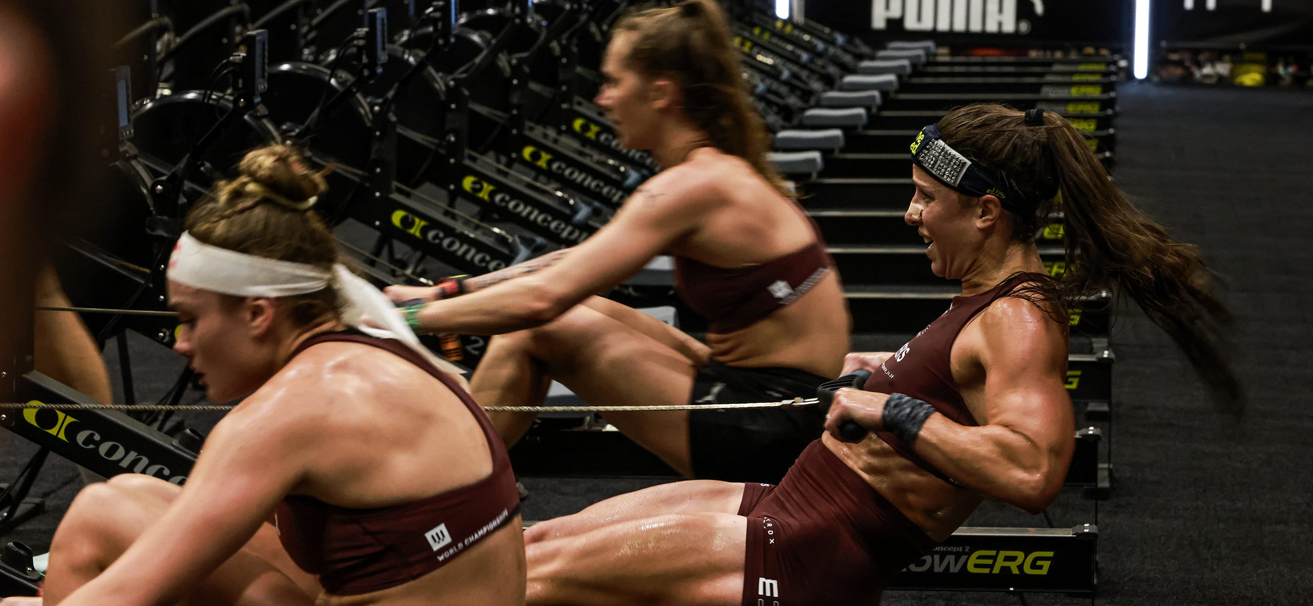 Female athletes intensely rowing on Concept2 erg machines during a fitness competition, showcasing strength, endurance, and focus in a high-performance training environment.
