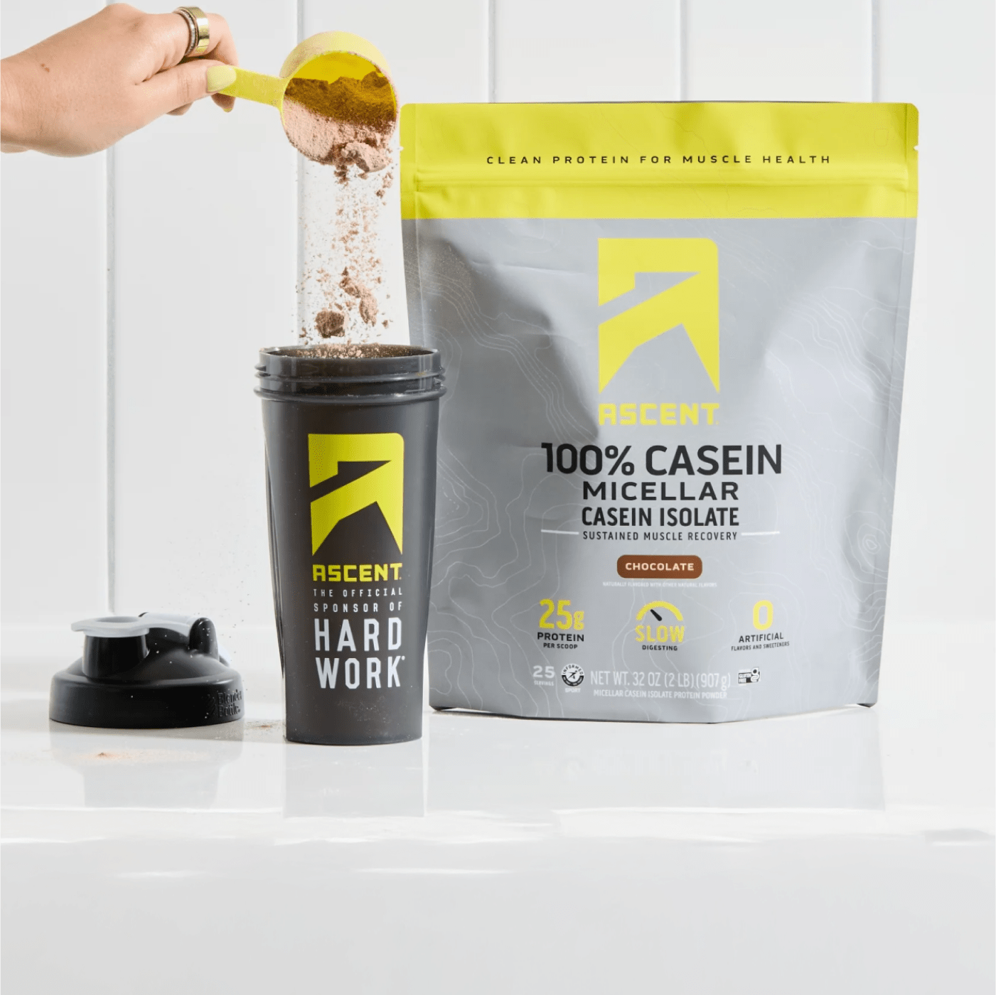 Ascent 100% Casein powder being scooped into shaker bottle next to large pouch