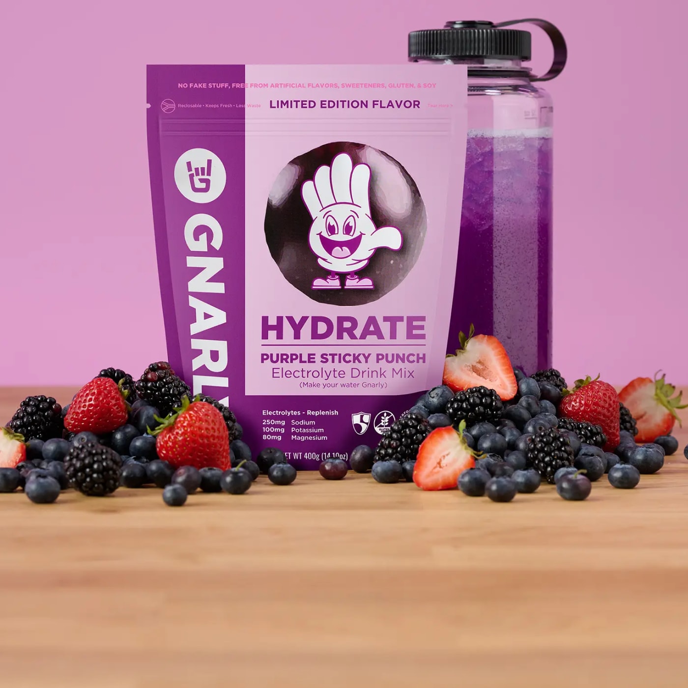 Limited edition Gnarly Hydrate Purple Sticky Punch electrolyte drink mix pouch with purple drink in a clear bottle and fresh berries on a wooden surface against a pink background