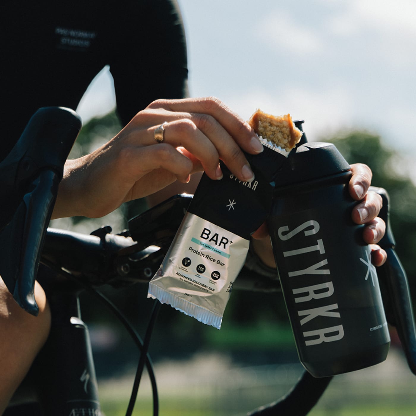 Cyclist unwrapping a STYRKR BAR+ protein rice bar while holding a black STYRKR water bottle, captured mid-ride on aero bars in a sunlit outdoor setting.