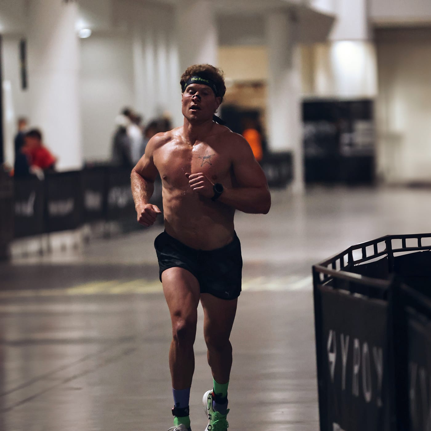 Hunter McIntyre running shirtless indoors during a HYROX event, wearing a green 'The Feed' headband, black shorts, and neon running shoes.