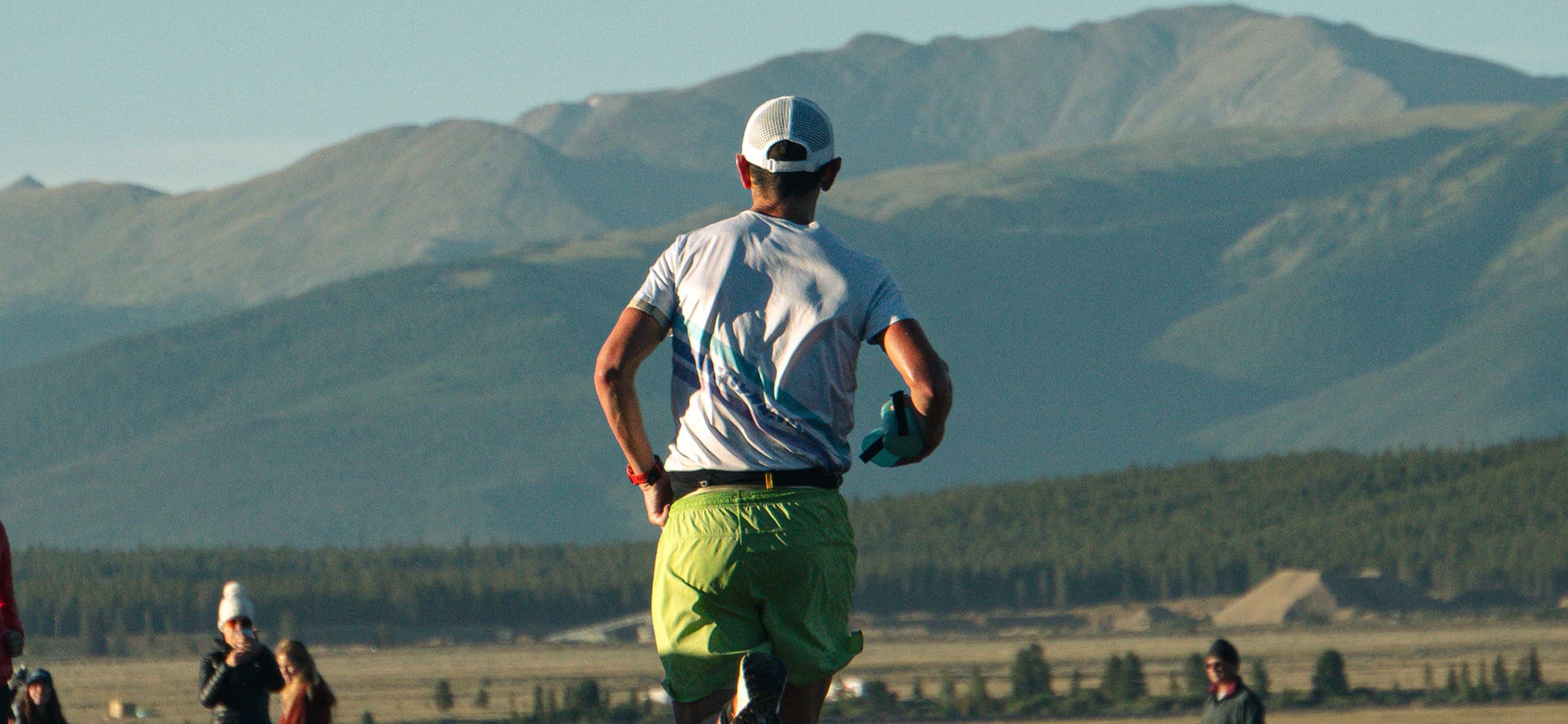 Trail runner David Roche runs toward the Colorado mountains during an endurance race, wearing a white cap, tech shirt, and green shorts.