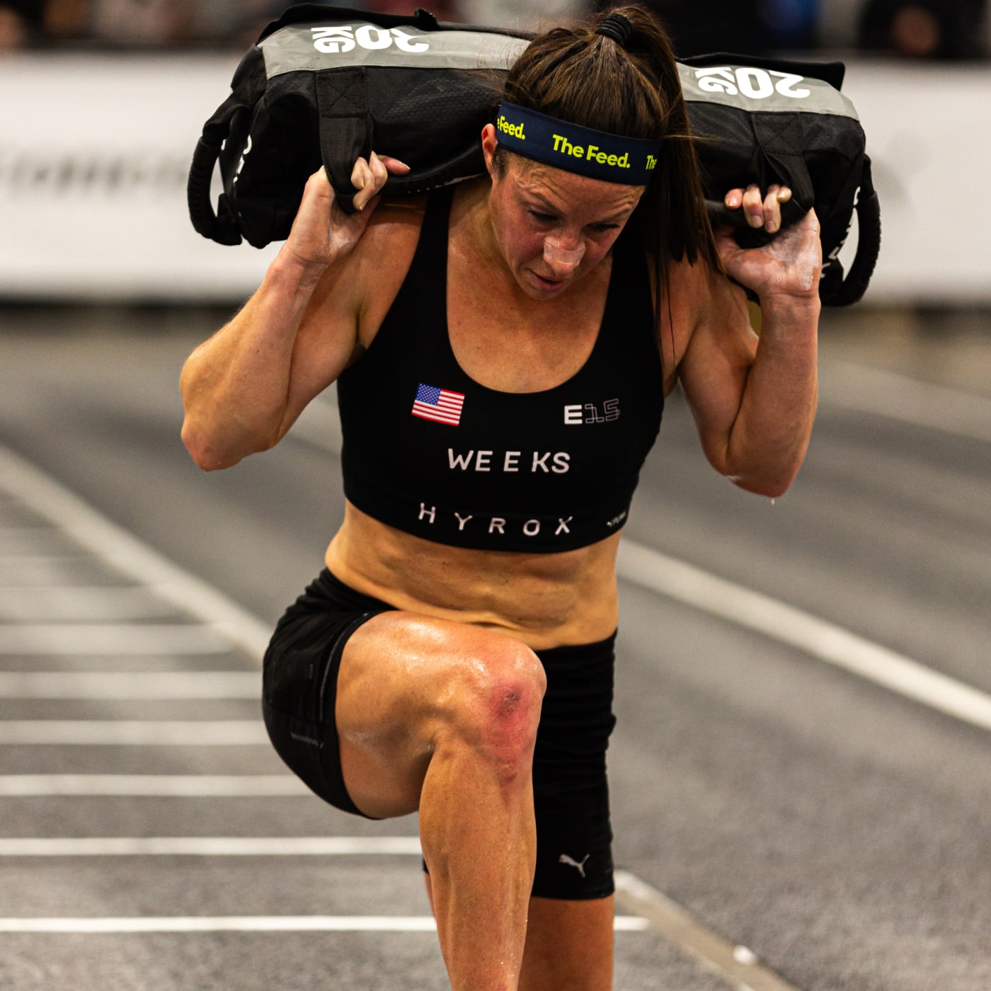 Female HYROX athlete performing weighted walking lunges with a sandbag across her shoulders, wearing The Feed headband and E15 race gear.