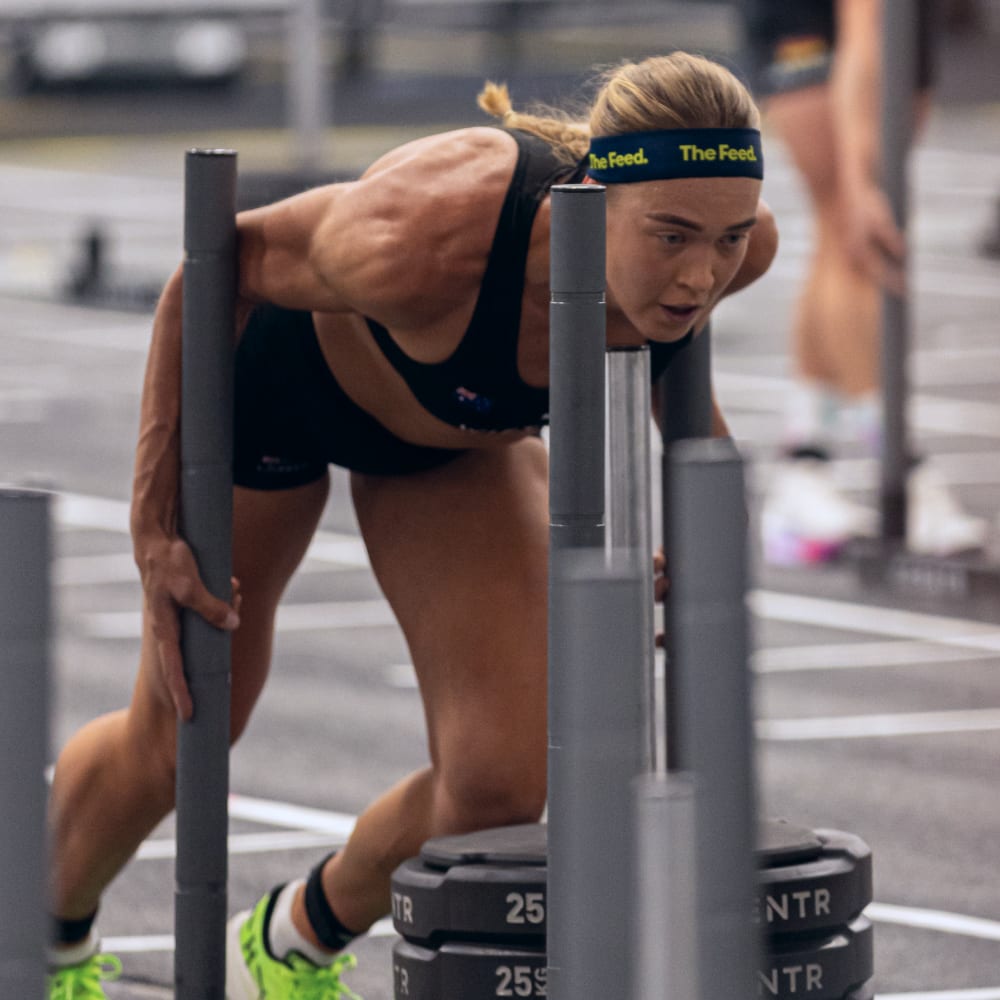 Focused HYROX athlete pushing a weighted sled during a competition, wearing The Feed headband and black training gear.