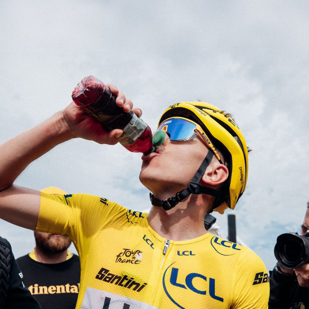 Tour de France champion Tadej Pogačar wearing the yellow jersey drinks Enervit Magic Cherry recovery drink after a stage finish, highlighting post-race recovery and performance nutrition.