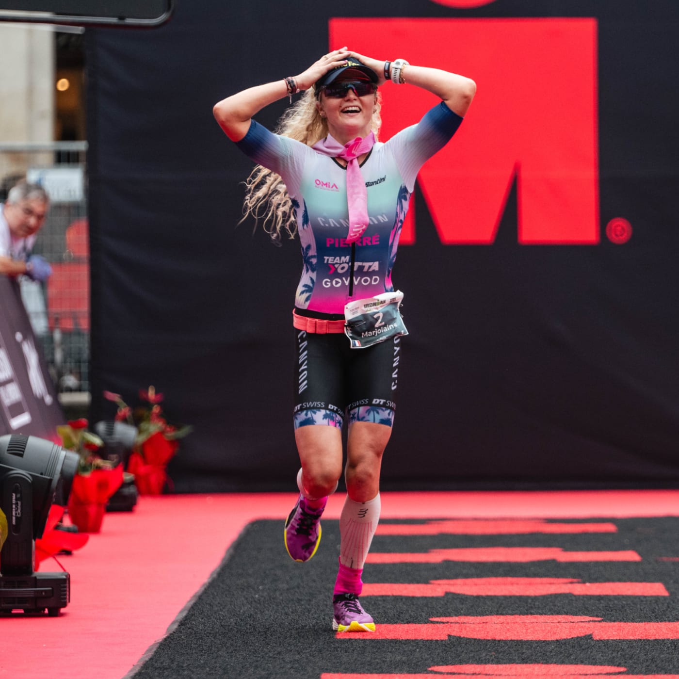 Professional triathlete Marjolaine Pierre crossing the Ironman finish line, smiling with hands on head in celebration, wearing Canyon team race kit and pink accessories.