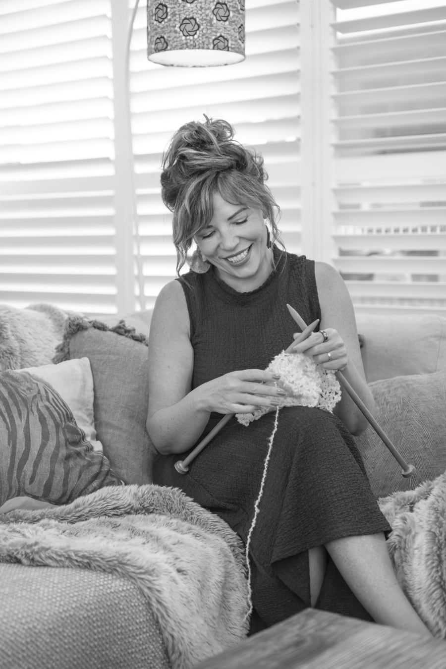 Dr Kirstey Holland, photographed in black and white, seated on a couch knitting and laughing