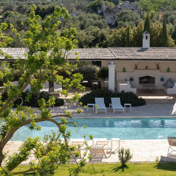 Views across the pool to the extensive, shaded communal area with fireplace, a lounge area, a dining table and a pool house.