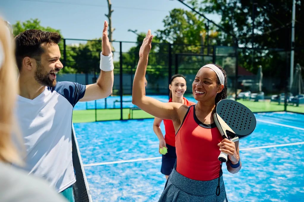 Padel players high-fiving on an enclosed court, showcasing the social and teamwork aspects of the sport. Padel players high-fiving on an enclosed court, showcasing the social and teamwork aspects of the sport.