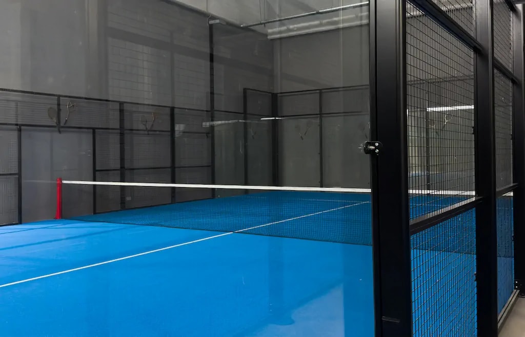 A modern indoor padel court with a blue playing surface, glass walls, and metal mesh. A modern indoor padel court with a blue playing surface, glass walls, and metal mesh.
