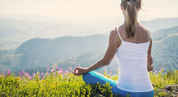 Donna in canottiera bianca e pantaloni blu seduta su una montagna erbosa a meditare dopo la riduzione del cappuccio clitorideo.