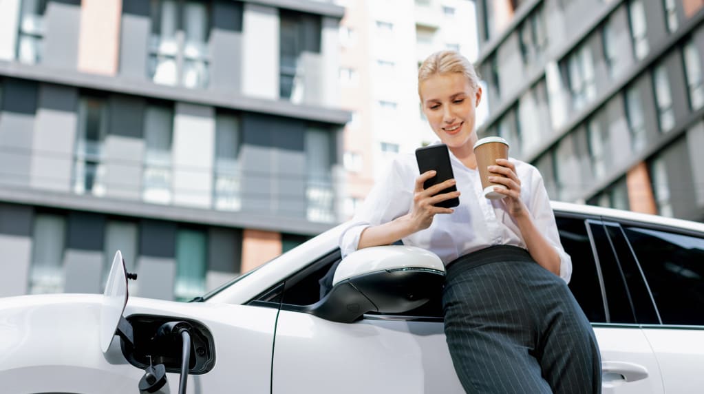 Woman with coffee near car charging station at an apartment