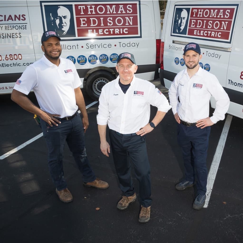 Three Thomas Edison Electric technicians stand with their work trucks