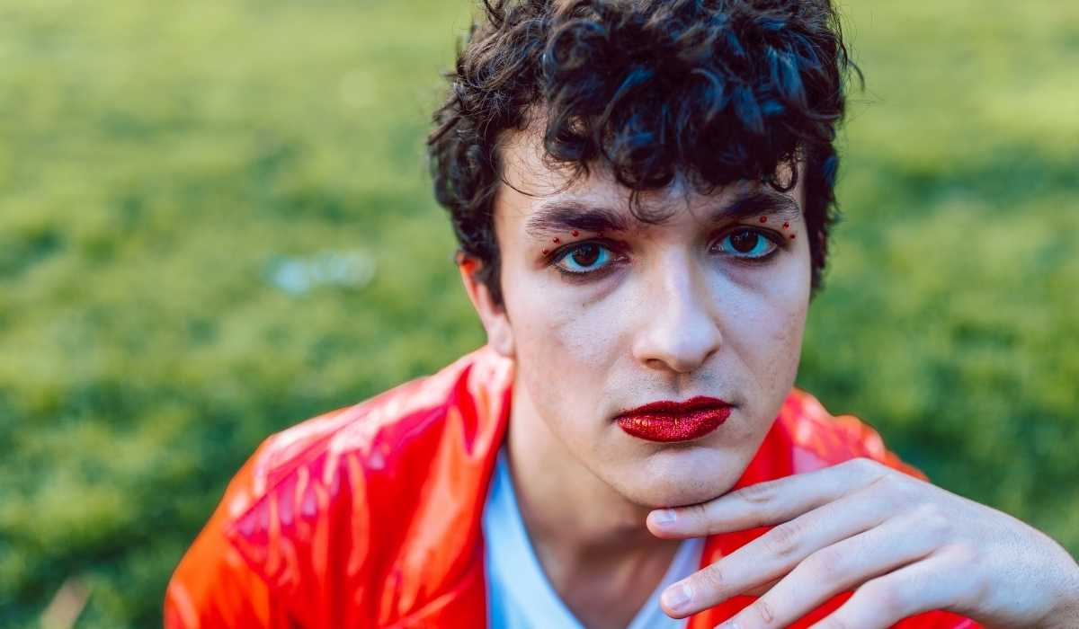 A young man in an orange jacket posing in the grass.