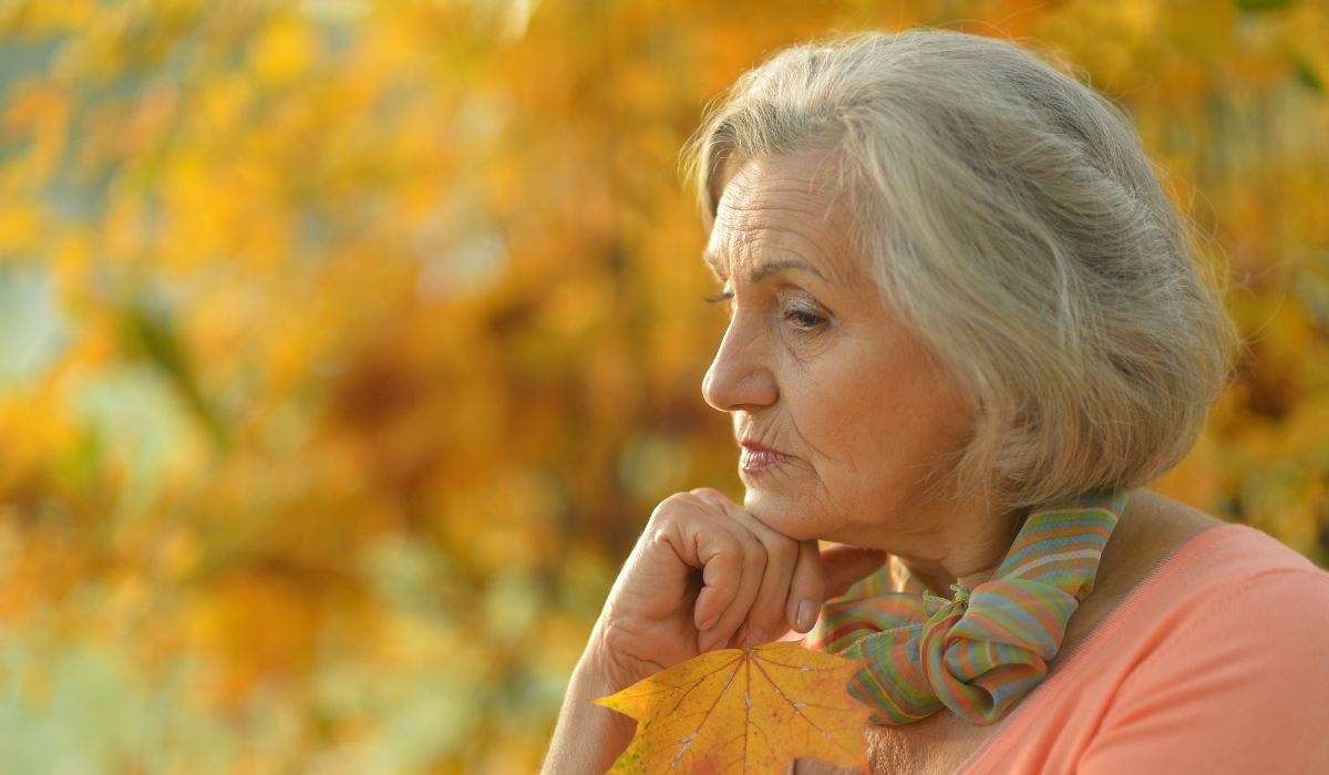 An elderly woman with gray hair holds a maple leaf, reflecting thoughtfully against the vibrant autumn foliage, embodying the essence of life transition therapy.