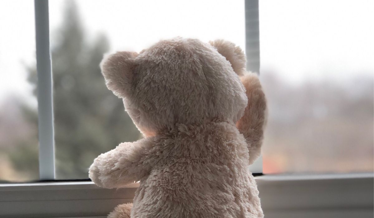 A light brown teddy bear sits on a windowsill, facing outside and touching the window with one paw, evoking memories of childhood trauma and longing.