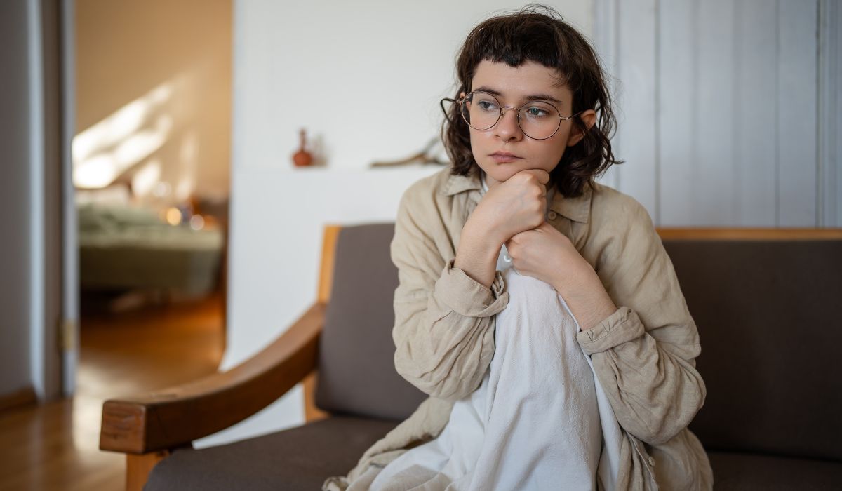 A thoughtful young woman with glasses sitting on a couch, resting her chin on her hand, looking contemplative.