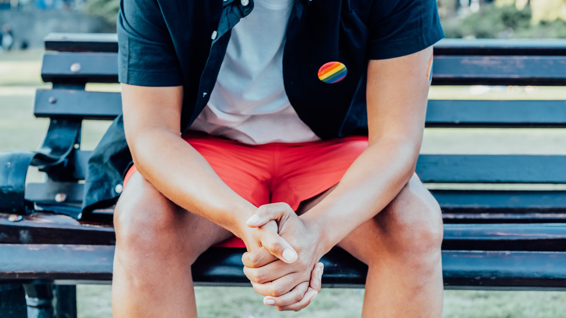 A person wearing a black shirt with a rainbow button and red shorts sits on a wooden bench, clasping their hands and looking down.