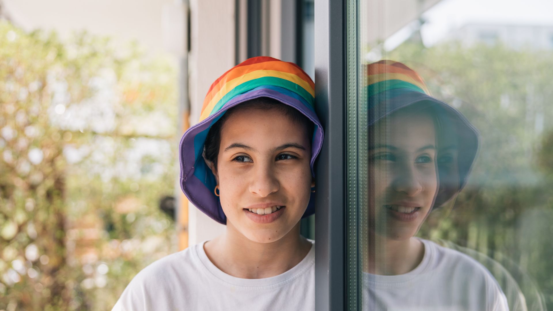 A person wearing a rainbow-colored hat and a white shirt is standing next to a glass wall, reflecting their image. Greenery is visible in the background.
