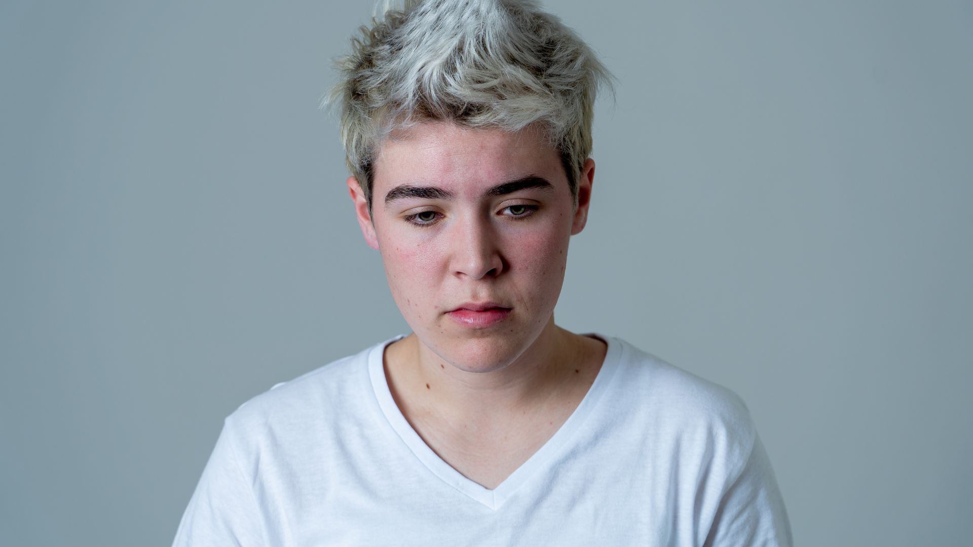 A person with short, light-colored hair wearing a white V-neck T-shirt stands against a plain background, looking down with a neutral expression.