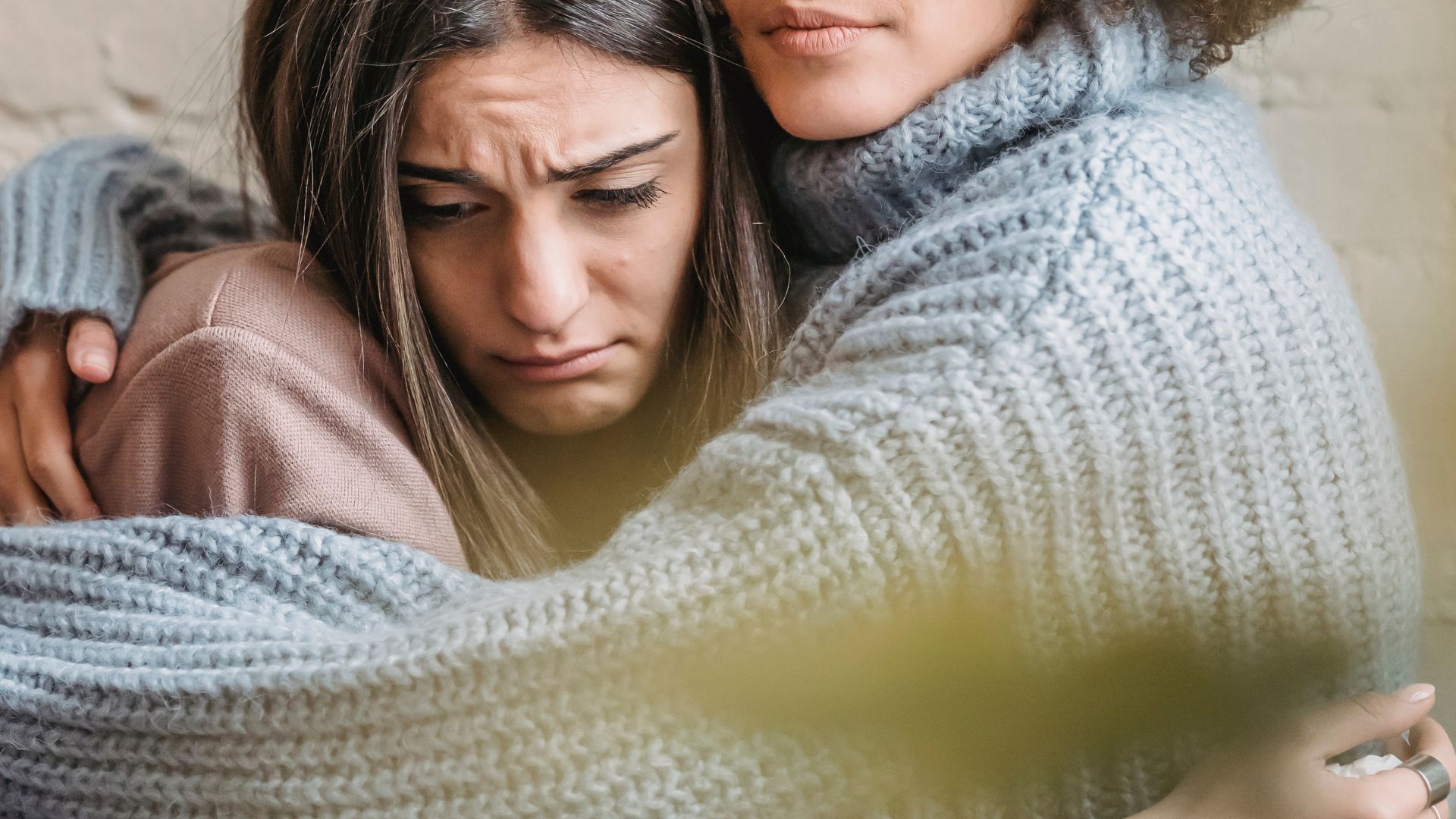 A person with long hair appears upset and is being consoled by another person wearing a gray sweater, who has their arm around them.
