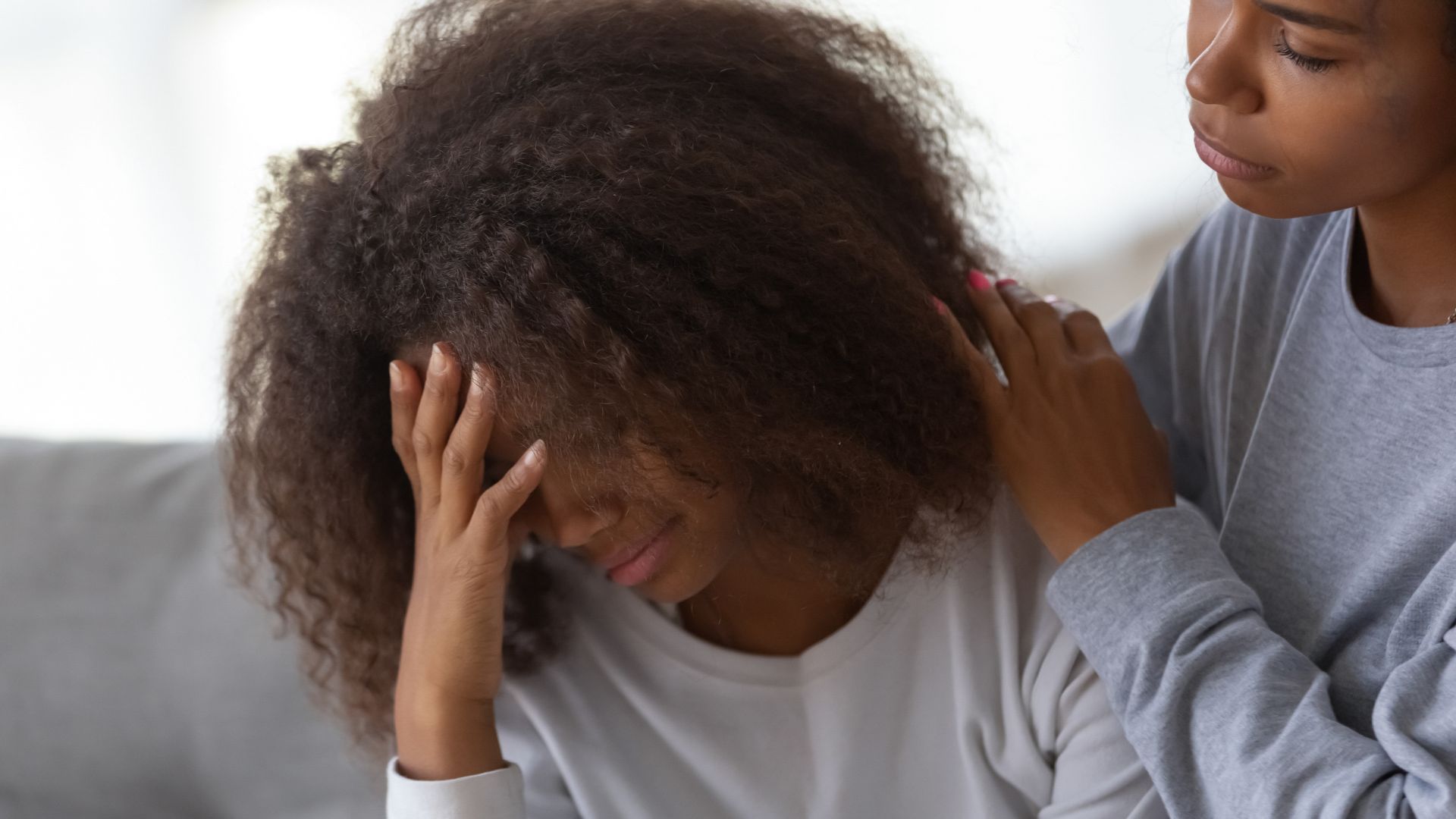A person with curly hair holds their head in their hand, looking distressed. Another person places a comforting hand on their shoulder.
