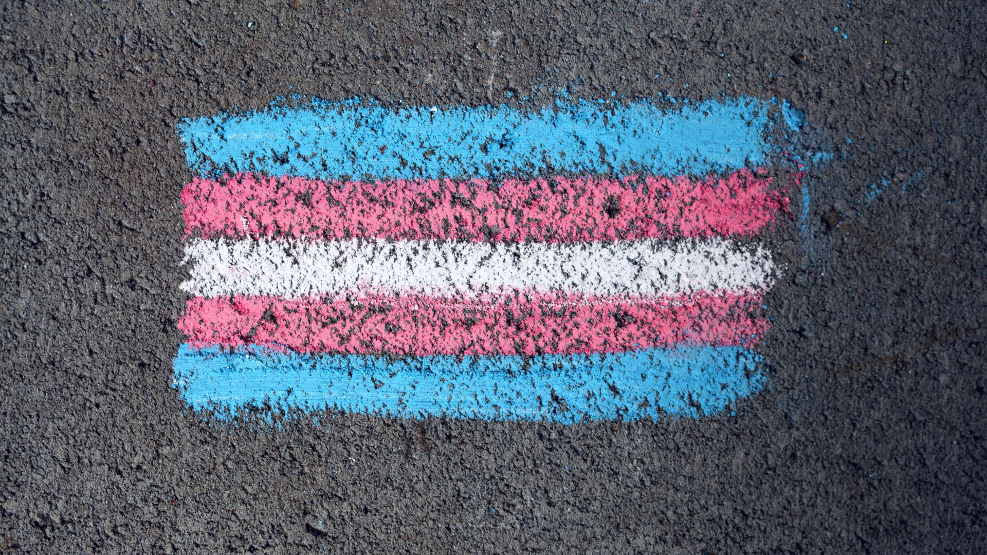 A transgender pride flag drawn with chalk on a rough, asphalt surface. The flag has blue, pink, and white stripes.