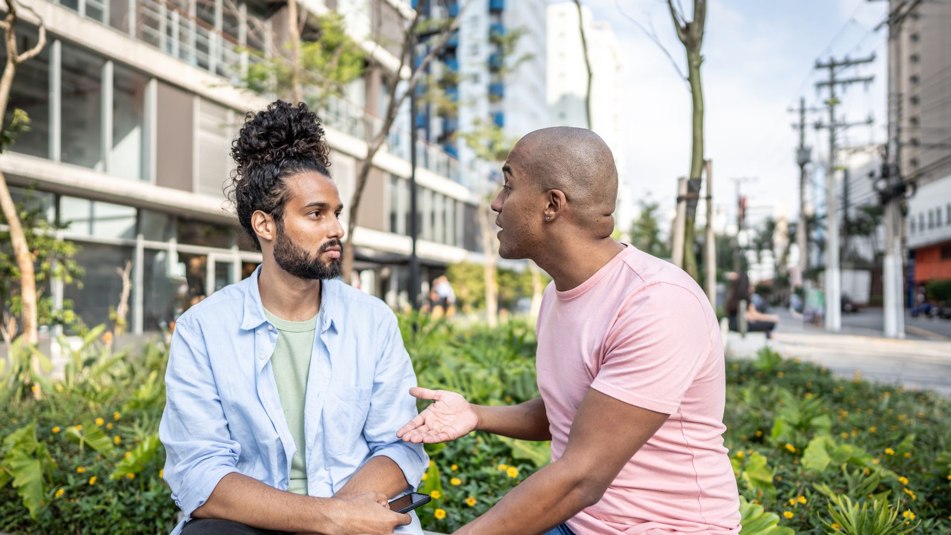 Two men sitting and talking on a bench near greenery in an urban setting on a sunny day, perhaps discussing enmeshment or trauma.