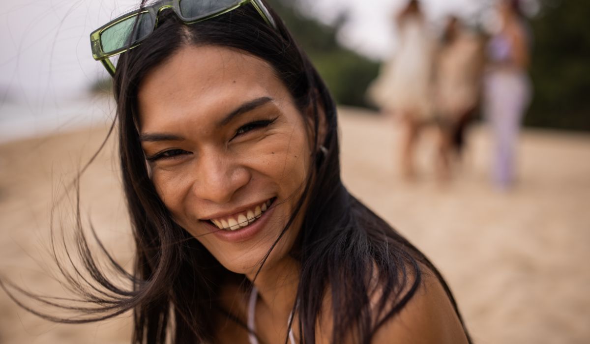 A person with long hair and sunglasses on their head smiles at the camera on a beach. People are visible in the blurred background.
