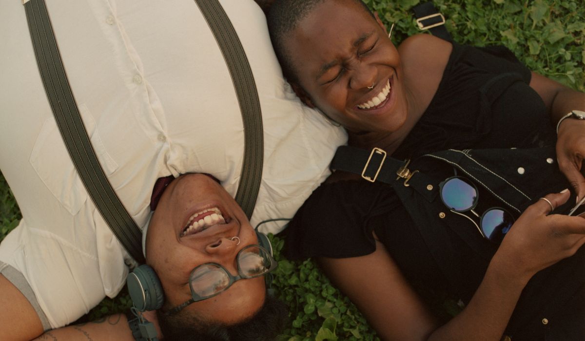 Two people lie on the grass, embodying the essence of a Queerplatonic Relationship. One smiles with headphones and glasses, while the other sports sunglasses hooked onto their overalls, both relaxed and content in each other's presence.
